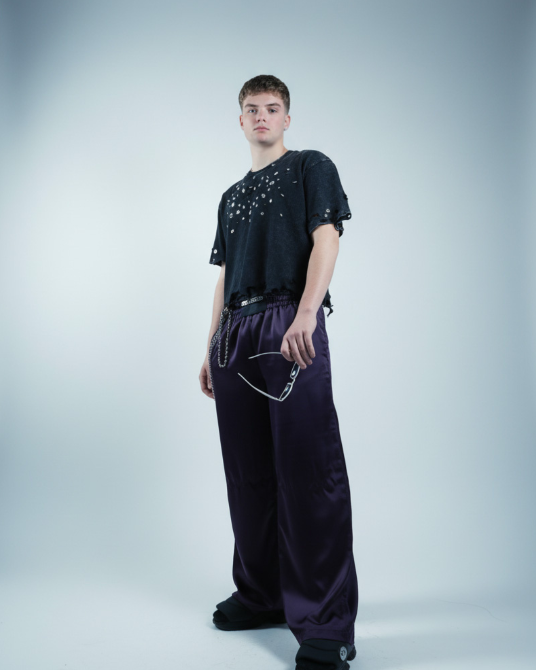 Farmhaus acid tears t-shirt. Model wearing a black embellished t-shirt woth studs and crystal and purple pants on a light gray background