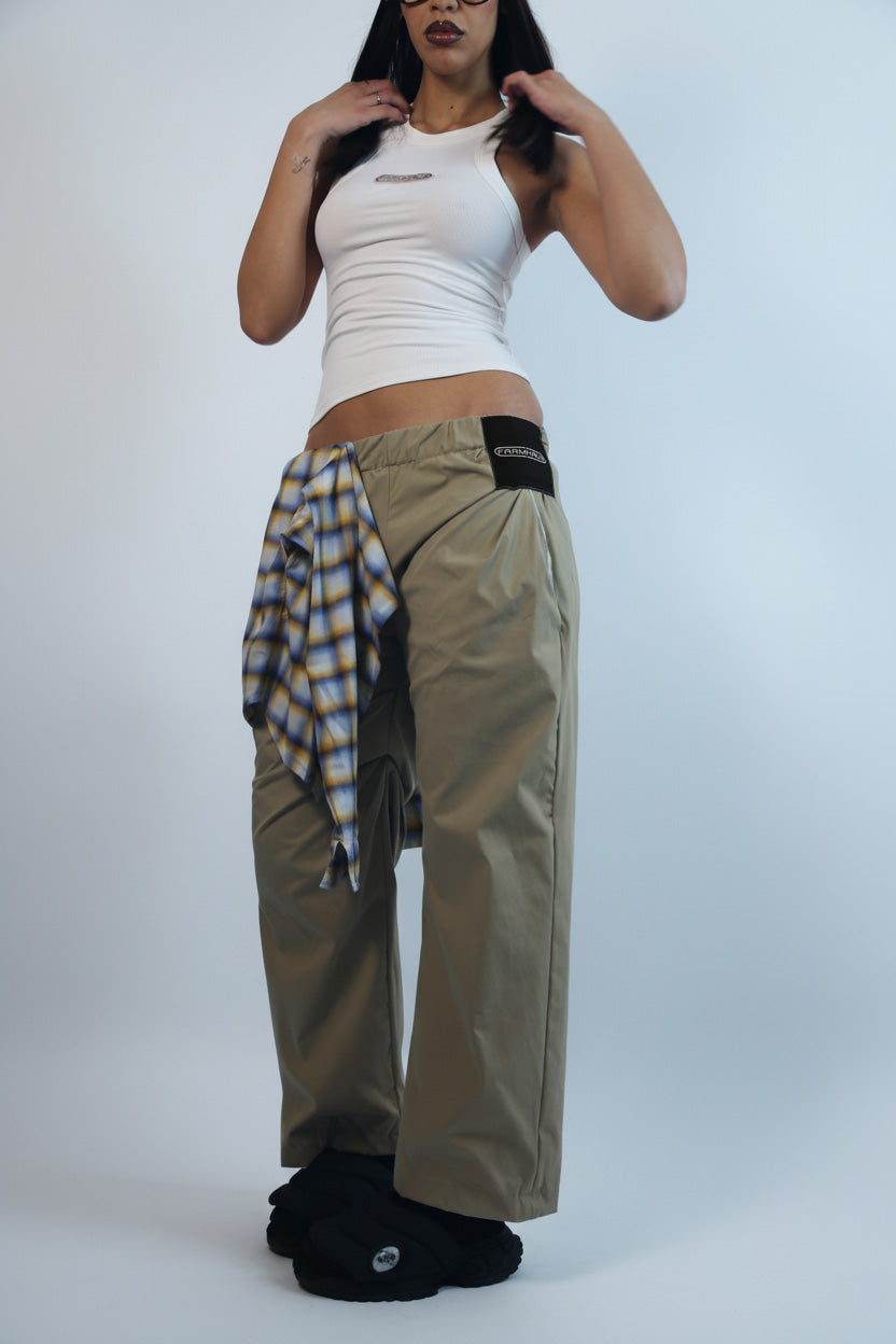 Person wearing a white tank top and beige pants with a plaid shirt over one shoulder on a white background