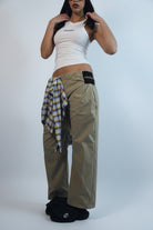 Person wearing a white tank top and beige pants with a plaid shirt over one shoulder on a white background