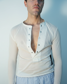 Man wearing a white long-sleeve shirt with a deep neckline and light blue pants against a plain background