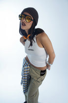 Person wearing a white tank top, beige pants, and a brown knit hat with sunglasses on a white background