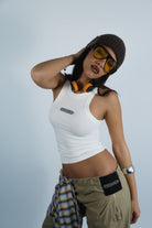 Person wearing a white tank top, brown pants, and a beanie with sunglasses on a light gray background