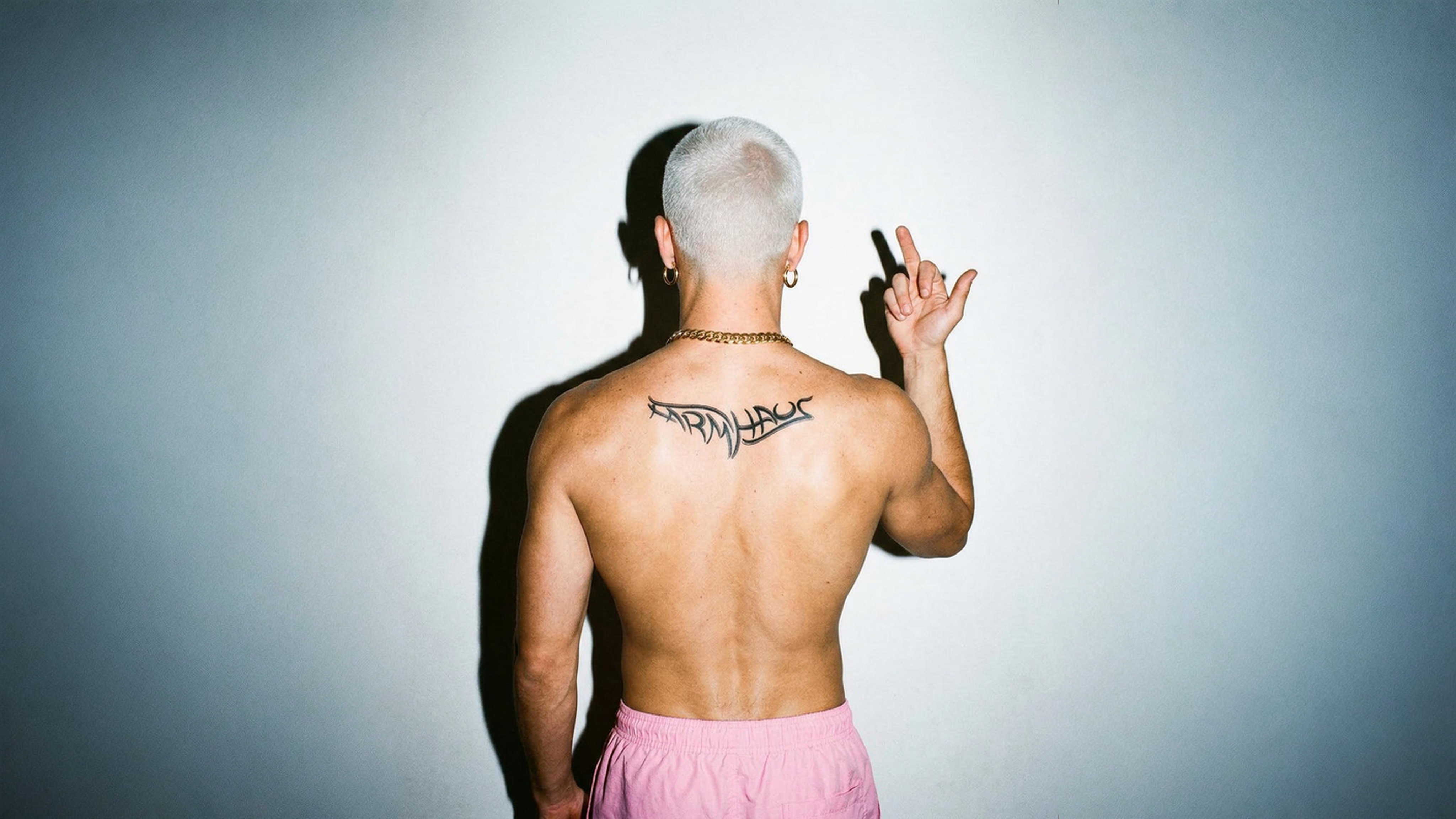 Person with shaved head and back tattoo making a peace sign against a gray background